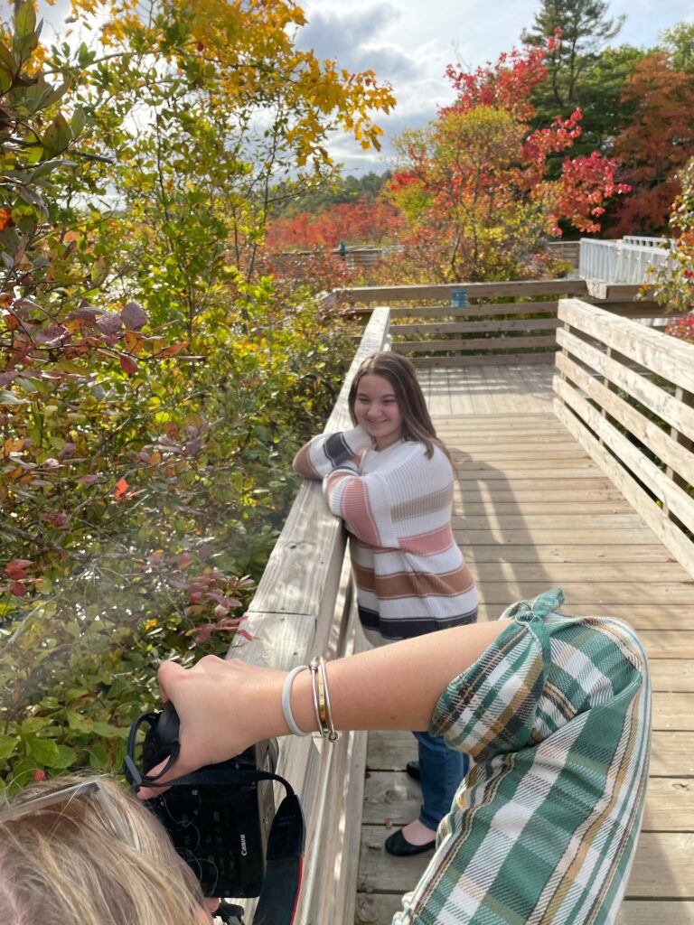 Photographer taking photos of high school senior on boardwalk in front of fall foliage.
