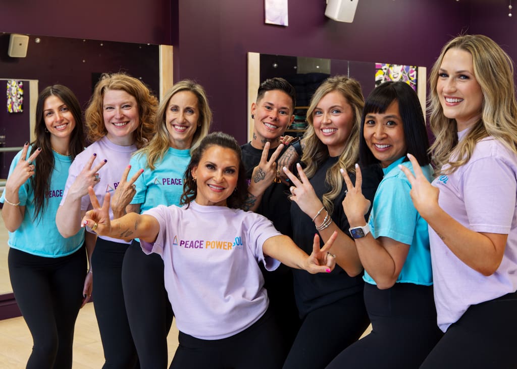 Group of women fitness instructors showing peace signs while smiling in branded t-shirts.