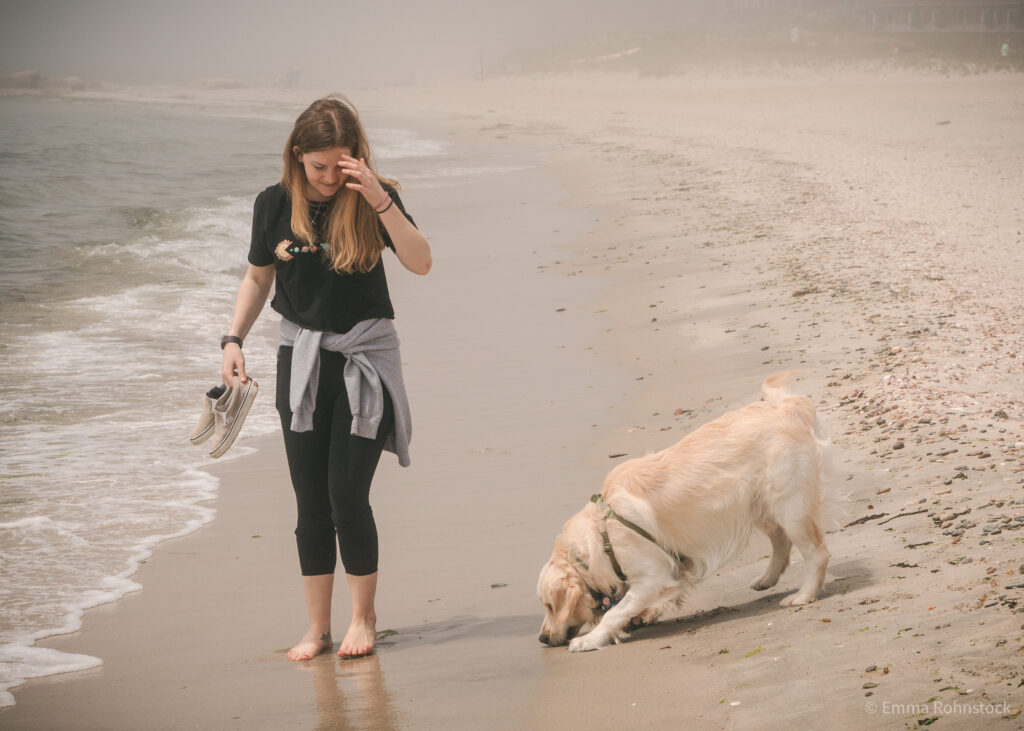 Woman holding shoes and walking along ocean waves with golden retriever.