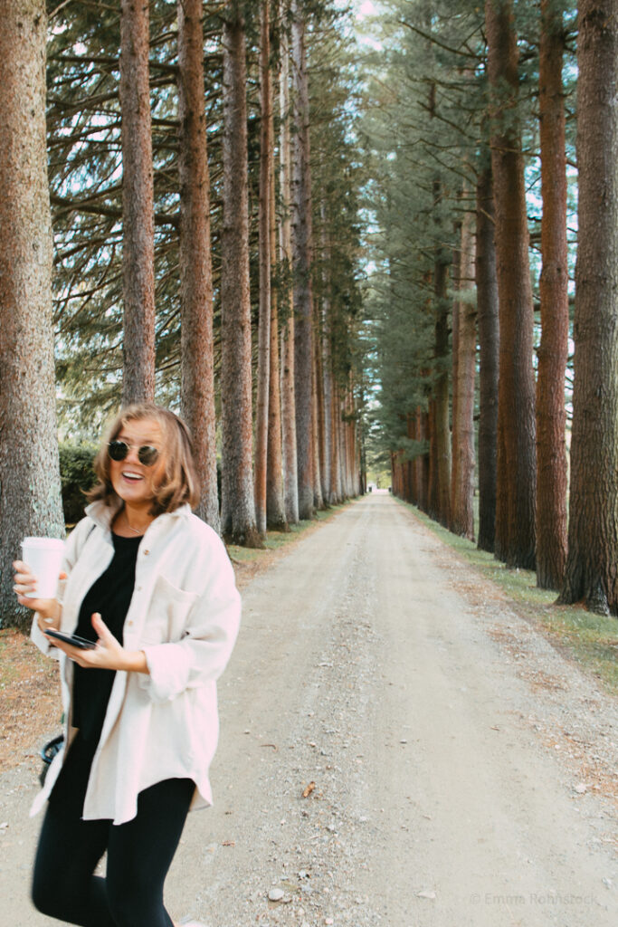 Woman smiling with a coffee in hand among rows of pine trees on a dirt path.