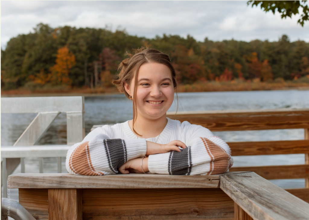 Girl smiling on boardwalk in front of water and fall foliage.