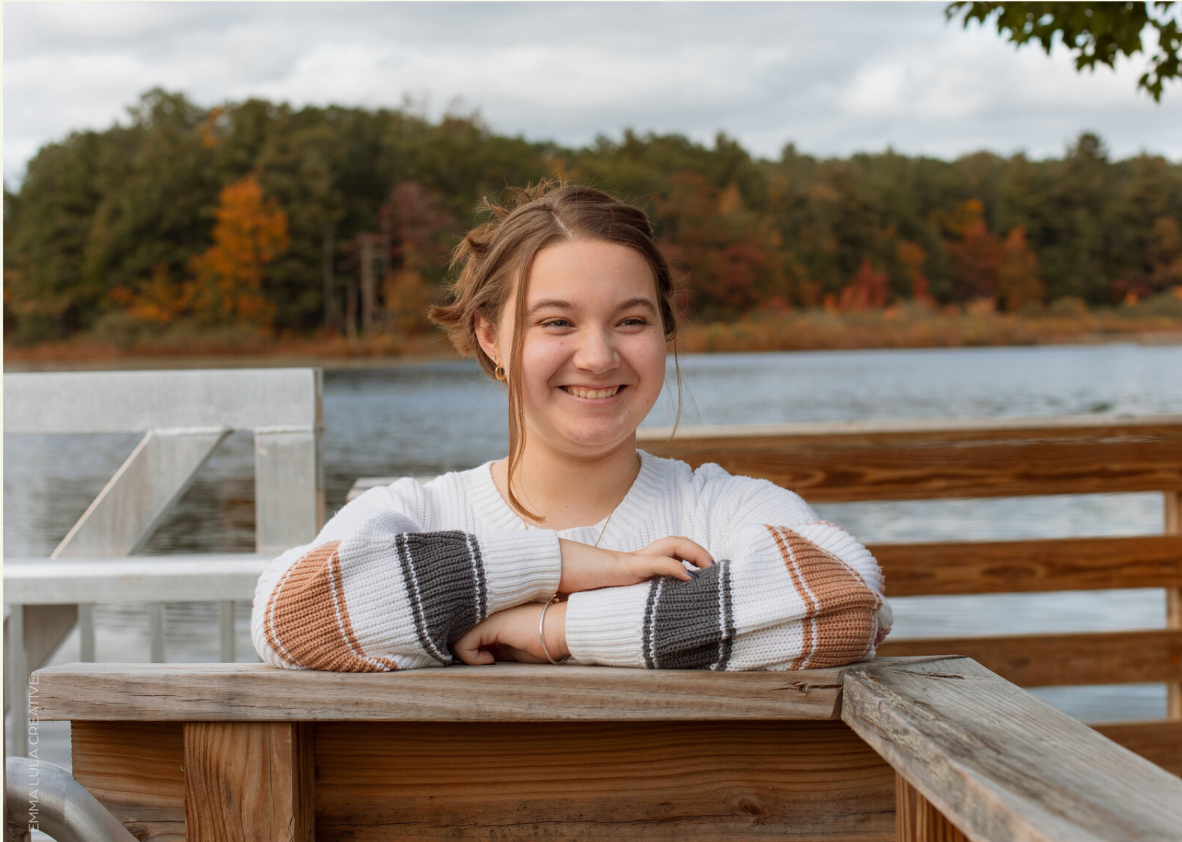 Girl smiling on boardwalk in front of water and fall foliage.